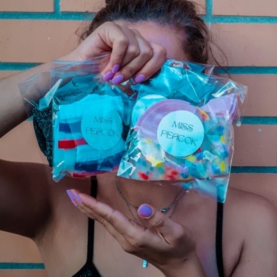 Pessoa segura dois sacos plásticos com etiquetas 'MISS PEACOCK' e tecidos coloridos.