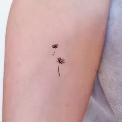 Tatuagem pequena de duas flores de dente-de-leão em tinta preta no braço