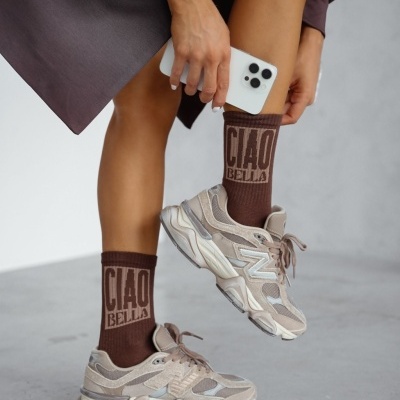 Meias castanhas com texto CIAO BELLA, ténis bege e cinza, telemóvel branco e roupa castanha escura.