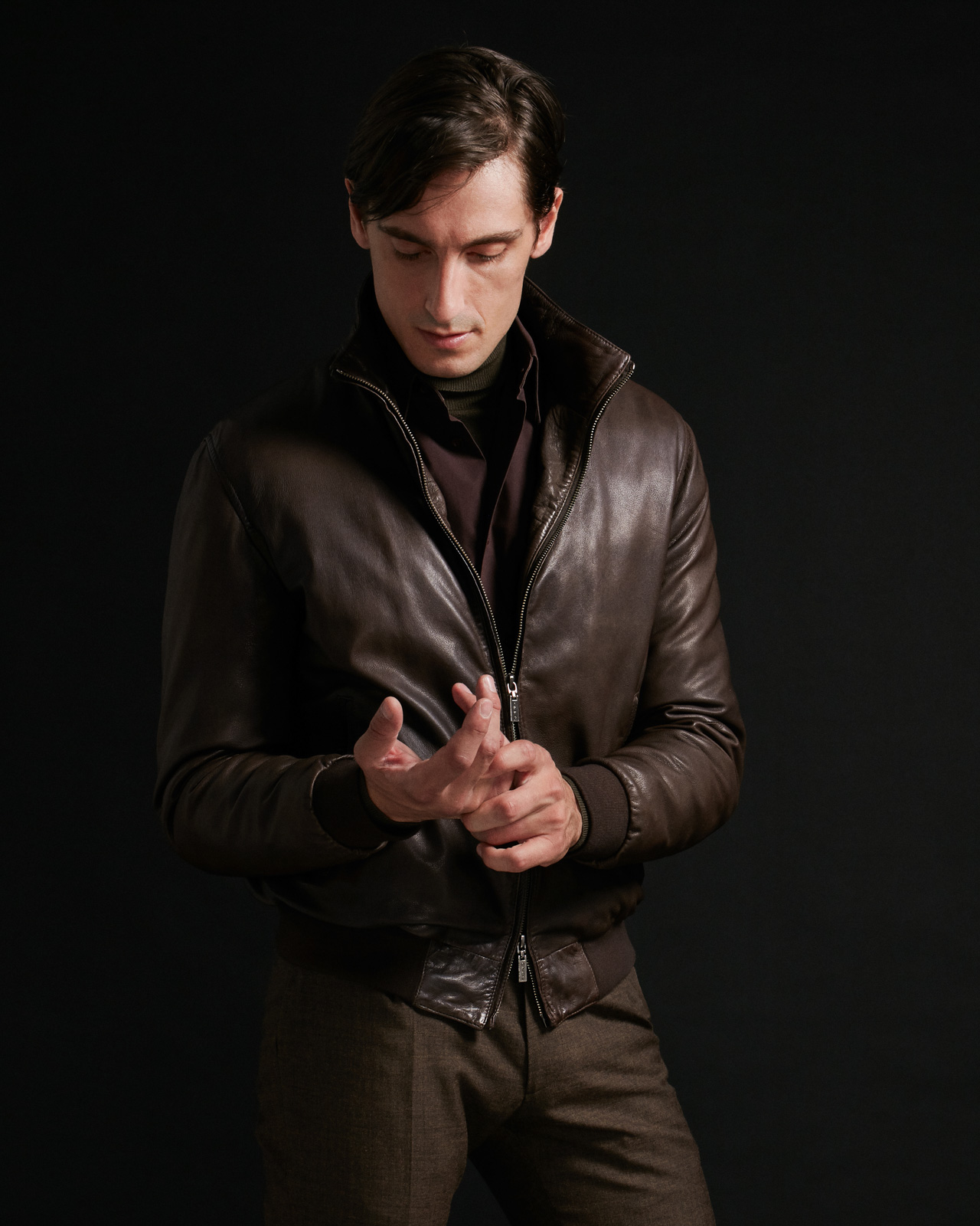 Homem com casaco de cabedal castanho escuro e calças castanhas frente a fundo preto