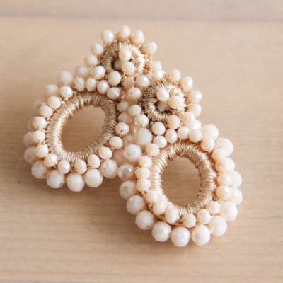 Brincos ovais bege com contas e fios dourados sobre superfície de madeira