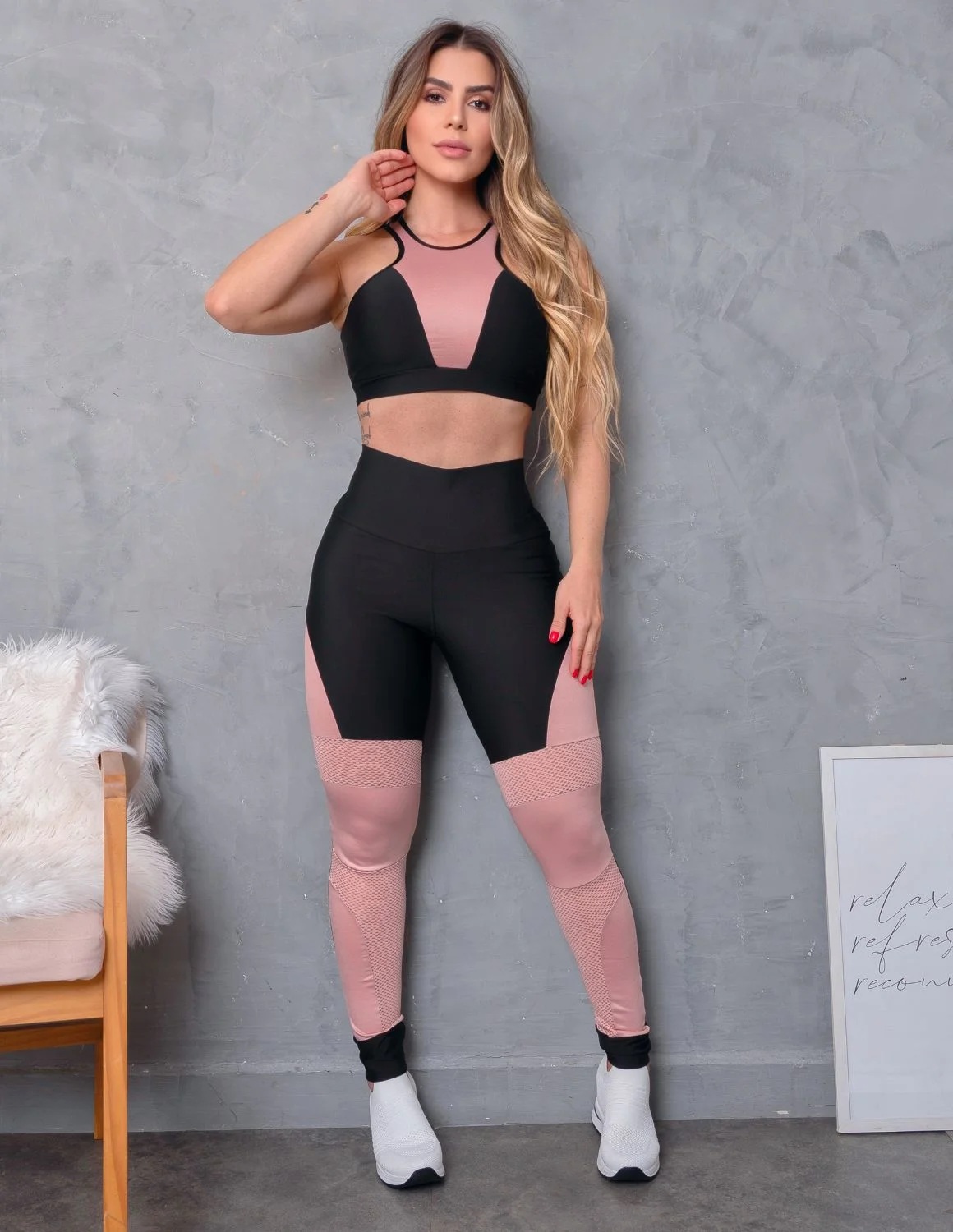 Look Feminino com Top sem Bojo e Calça com Recortes em Tela e Cirrê Mulher vestindo conjunto desportivo preto e rosa posando contra parede cinza com cadeira e quadro ao lado