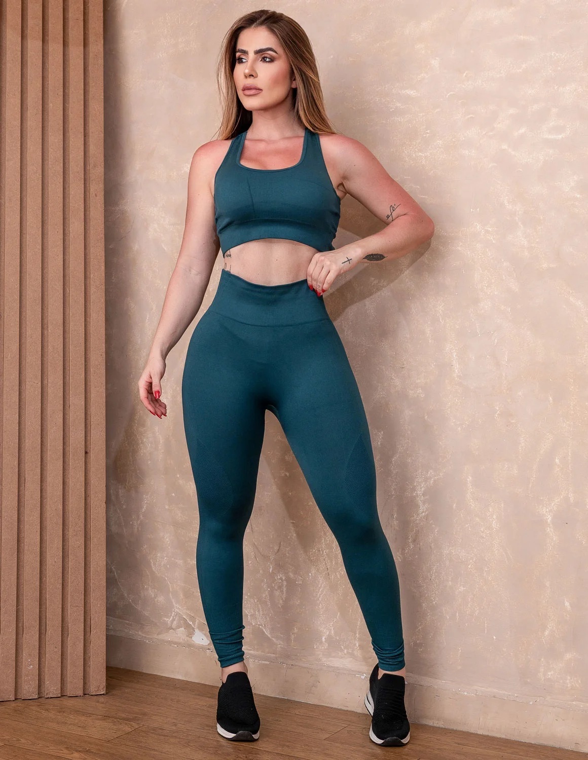 Conjunto Seamless de Poliamida com Calça Cintura Alta e Cropped Trançado Mulher com conjunto desportivo azul petróleo e sapatilhas pretas, em interior com parede bege.