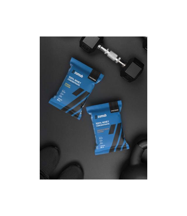 Whey (Saquetas) Duas embalagens azuis de suplemento proteico Zumub 100% Whey Concentrate junto a equipamento de ginásio preto.