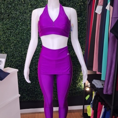 Conjunto desportivo feminino roxo composto por top halter e leggings com saia incorporada, em manequim branco sobre base preta, frente a parede verde artificial e roupas penduradas.