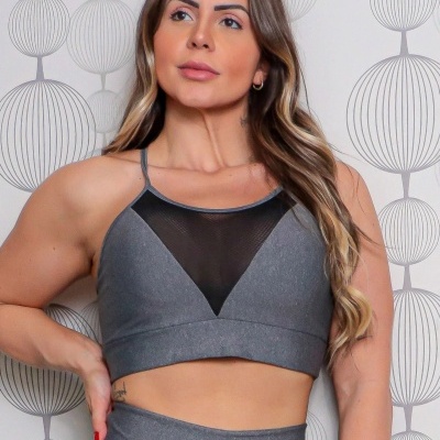 Top desportivo cinzento com detalhe em tela preta usado por mulher em fundo com padrão geométrico.