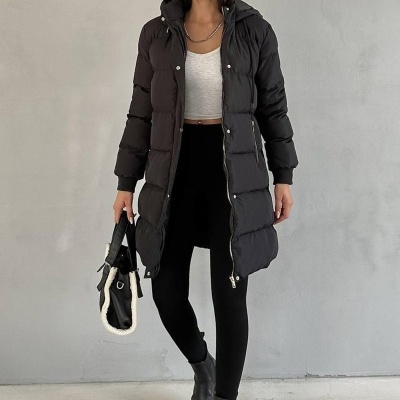 Mulher com casaco acolchoado preto, botas e boné cinza em fundo cinza.