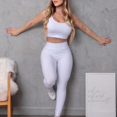 Mulher com roupa desportiva branca encostada a parede cinza com cadeira com almofada branca e quadro com texto.