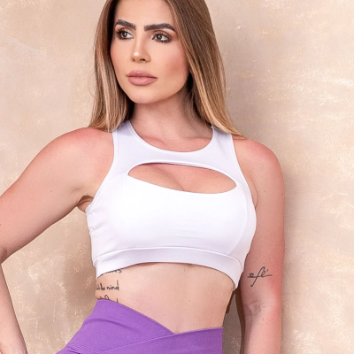 Mulher de top desportivo branco com recorte e calças de treino roxas frente a fundo bege