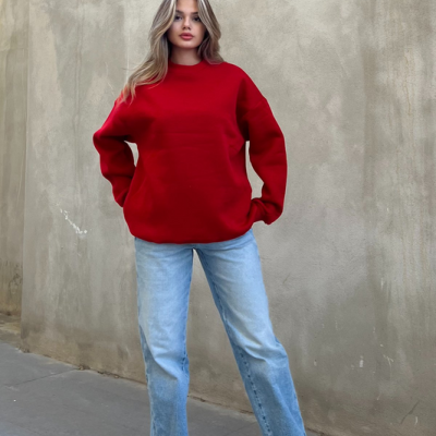 Mulher com camisola vermelha, jeans azul claro e ténis brancos em fundo de parede bege texturada.