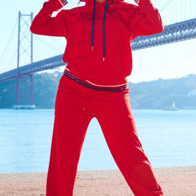 Mulher veste conjunto desportivo vermelho com capuz e calças largas, com ténis brancos, junto a uma ponte sobre o rio.