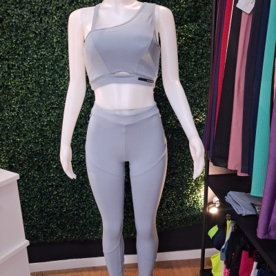 Conjunto desportivo feminino cinzento claro com top assimétrico e leggings exibido em manequim branco