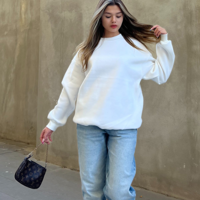 Mulher com sweatshirt branca e jeans azuis caminhando numa rua com mala preta