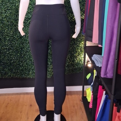 Leggings pretas justas em manequim de costas, parede verde artificial e prateleira com roupas coloridas ao lado