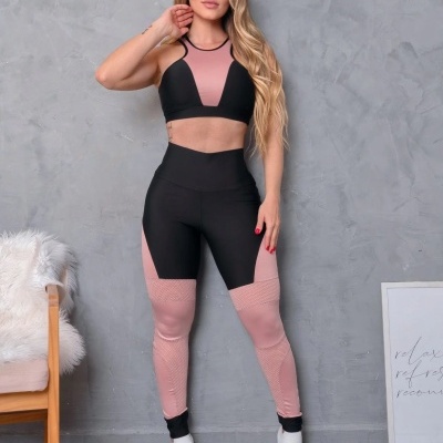 Mulher vestindo conjunto desportivo preto e rosa posando contra parede cinza com cadeira e quadro ao lado
