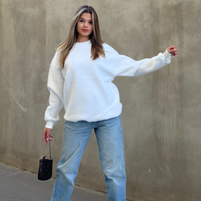 Mulher com sweatshirt branca, jeans azuis, ténis brancos e mala preta junto a parede cinza
