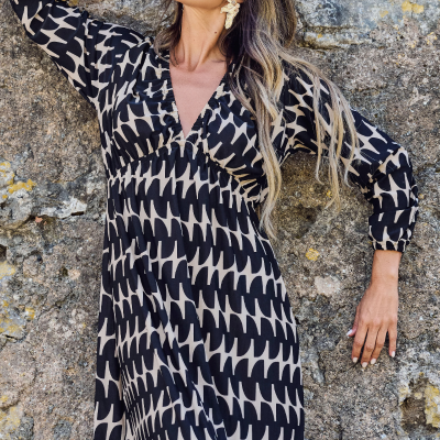 Vestido longo preto e bege com padrão geométrico usado por mulher com óculos e brincos perto de parede de pedra