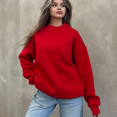 Mulher usa camisola vermelha de manga comprida e jeans claros junto a uma parede bege