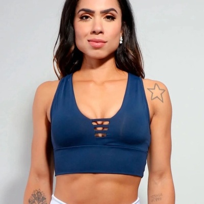 Top desportivo azul escuro com detalhes em tiras, usado por mulher com tatuagens e calças brancas, fundo cinza claro