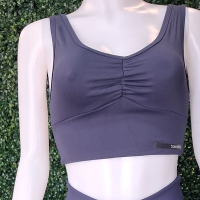 Top desportivo cinza escuro com franzido central em manequim branco junto a parede de folhas verdes.