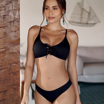 Mulher vestindo biquíni preto de tecido liso com cordão frontal, brincos azuis, em ambiente interior com sofá cinza e azulejos azuis no chão