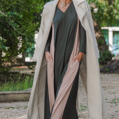 Mulher com vestido verde escuro e rosa, casaco bege, botas e óculos de sol ao ar livre