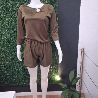 Macacão curto castanho com mangas três quartos em manequim branco, fundo com folhagem artificial e planta em vaso cinza