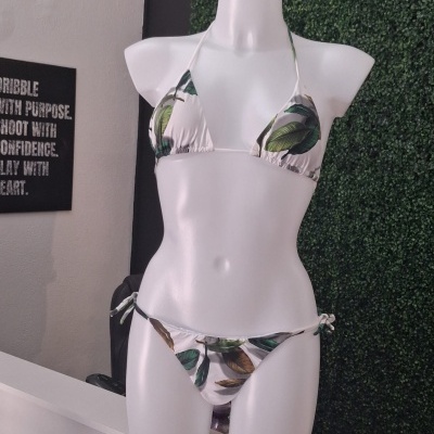 Biquíni branco com folhas verdes e castanhas, mostrado num manequim branco com fundo de parede verde artificial e parede branca com quadro negro.