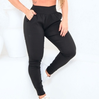 Calças pretas justas com bolsos e ténis brancos numa parede branca.