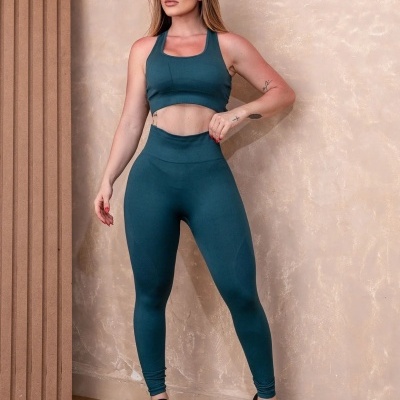 Mulher com conjunto desportivo azul petróleo e sapatilhas pretas, em interior com parede bege.