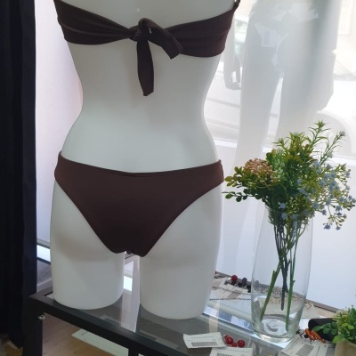 Biquíni castanho em manequim feminina, visto de costas, com vaso de flores ao lado sobre mesa de vidro