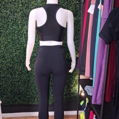 Manequim com roupa desportiva preta, top e leggings, com fundo de plantas artificiais e roupas coloridas ao lado.