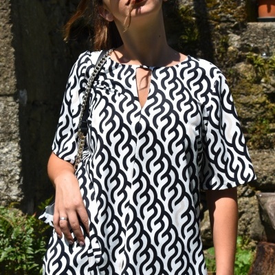 Pessoa de túnica branca com padrão preto em ambiente exterior com plantas e pedras