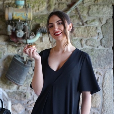 Mulher com vestido preto e cabelo apanhado, sorrindo num ambiente rústico com parede de pedras