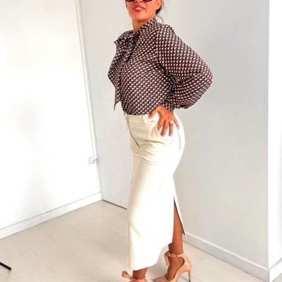 Mulher com blusa padrão geométrico, saia branca com fenda e sapatos bege num ambiente claro