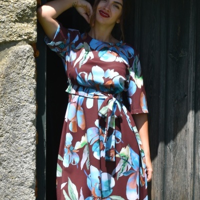 Vestido floral castanho com flores azuis e folhagem verde, com cinto, usado por mulher junto a parede de pedra e porta de madeira