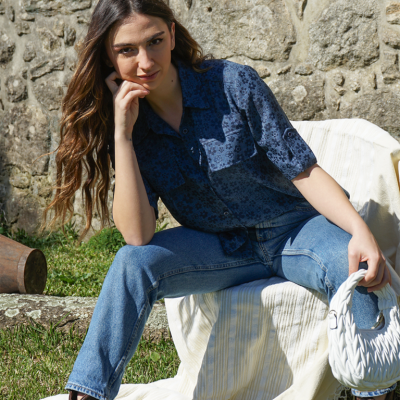 Mulher sentada com camisa azul estampada, calças de ganga e mala branca ao ar livre junto a parede de pedra