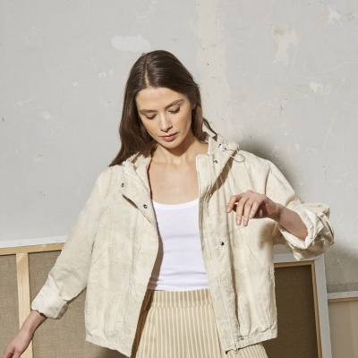 Casaco feminino bege claro com padrão texturizado e capuz, com zíper e botões, usado com top branco e saia riscada.