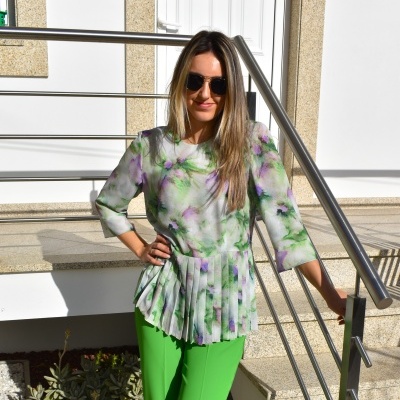 Mulher usando blusa floral branca com plissados e calças verdes encostada em corrimão