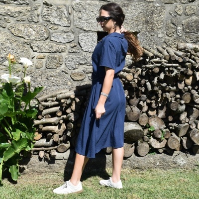 Mulher de vestido azul e ténis brancos junto a parede de pedra e lenha