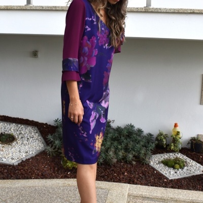 Mulher com vestido floral roxo e sapatos pretos em área exterior com jardim e muro branco