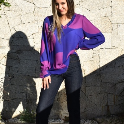 Mulher a usar blusa azul e roxa com padrão geométrico e jeans escuros em exterior com parede de pedra