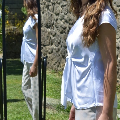 Mulher com camisa branca e calças bege com padrão de círculos diante de espelho ao ar livre