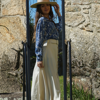 Mulher com chapéu de palha, blusa azul com padrão floral e saia branca reflectida em espelho exterior perto de parede de pedra