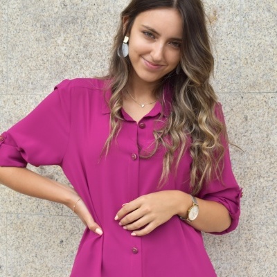 Mulher com camisa rosa forte de botões e mangas dobradas, parede cinza ao fundo