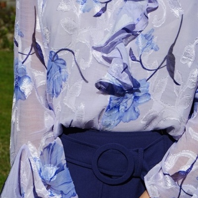 Blusa branca com padrão floral azul e folhos, cinto azul escuro na cintura.