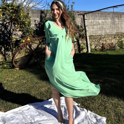Mulher a usar vestido verde claro fluido com mangas até ao cotovelo e gola em V, em jardim com relva, cobertor branco no chão.