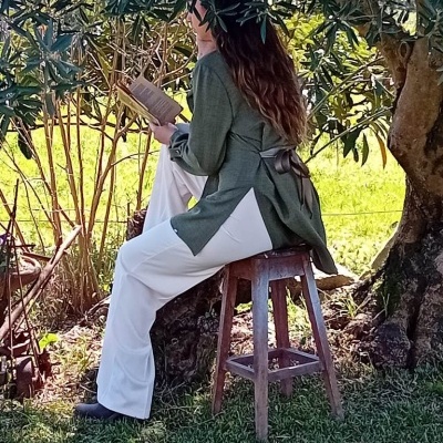 Mulher sentada num banco de madeira no jardim a ler um livro, com casaco verde, calças brancas e sapatos pretos