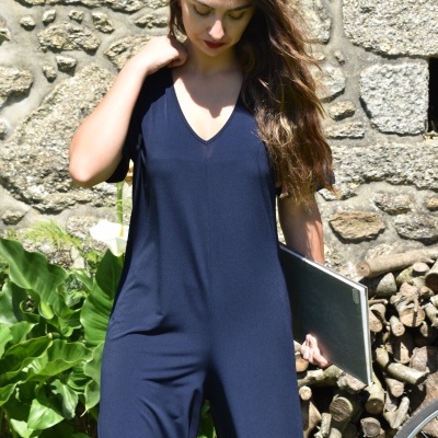 Mulher com macacão azul-escuro segurando caderno preto em frente a parede de pedra