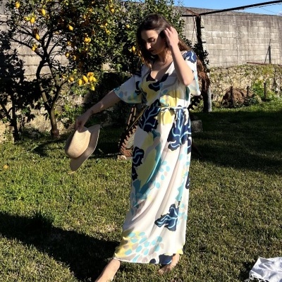 Mulher com vestido comprido floral azul e amarelo segurando chapéu de palha em jardim com árvore de limão